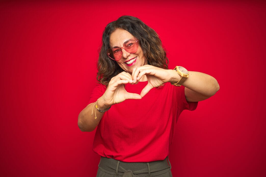 Middle age senior woman wearing cute heart shaped glasses over red isolated background smiling in love showing heart symbol and shape with hands. Romantic concept. | Meridian Source