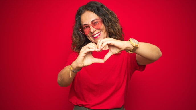 Middle age senior woman wearing cute heart shaped glasses over red isolated background smiling in love showing heart symbol and shape with hands. Romantic concept. | Meridian Source