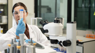 Female Scientist Examining Test Tube Sample in Modern Research Laboratory with Microscope | Meridian Source